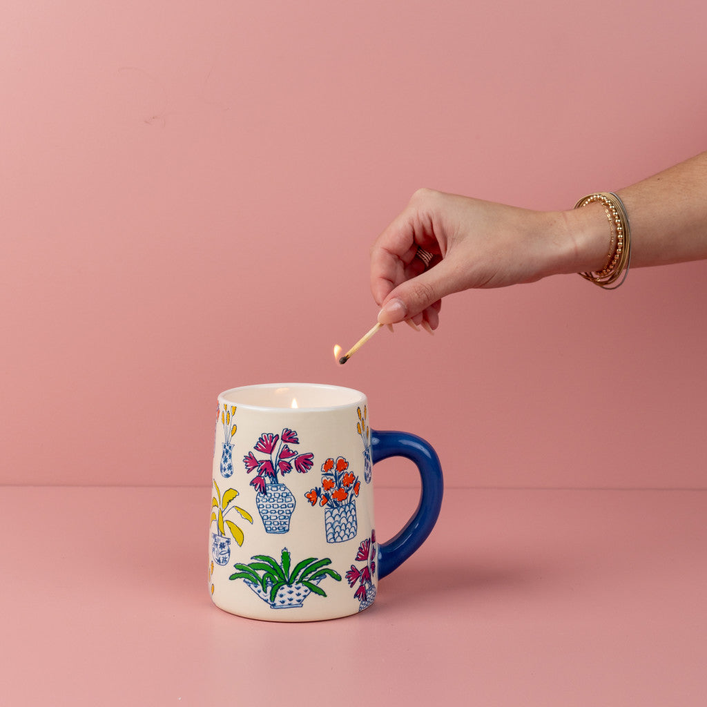 A hand with gold bangles lights a match above the Bridgewater Candle Company Sweet Grace Collection Candle #054 in a white mug with blue handle and potted plant designs—a must-have for any plant mom. Perfect for your favorite soy-blend wax candle.
