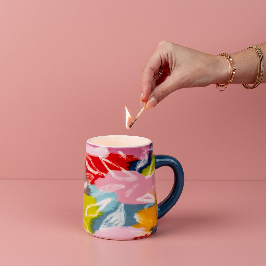 A hand lights a match to the Bridgewater Candle Company Sweet Grace Collection Candle #055, made from soy-blend wax in a colorful, abstract-patterned mug with a blue handle, set against a pink background.