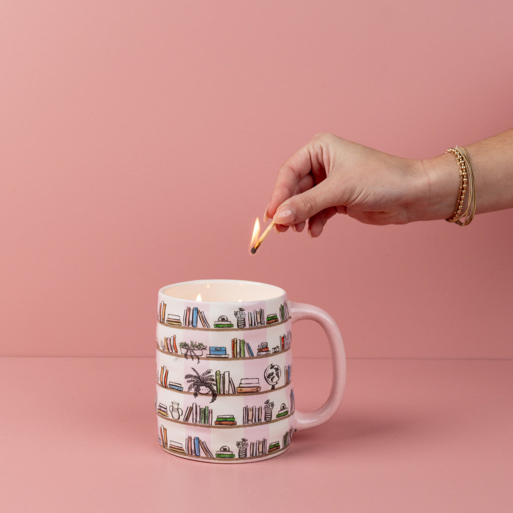 A hand lights the Bridgewater Candle Company Sweet Grace Collection Candle #056, set in a bookshelf-illustrated mug with a handle against a pink background. The candle’s premium soy-blend wax creates a cozy ambiance.