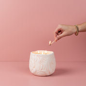 A hand with bracelets lights the Bridgewater Candle Company Sweet Grace Collection Candle #057—made of soy-blend wax with a white floral pattern—using a match, against a soft pink background for a touch of luxury home fragrance.