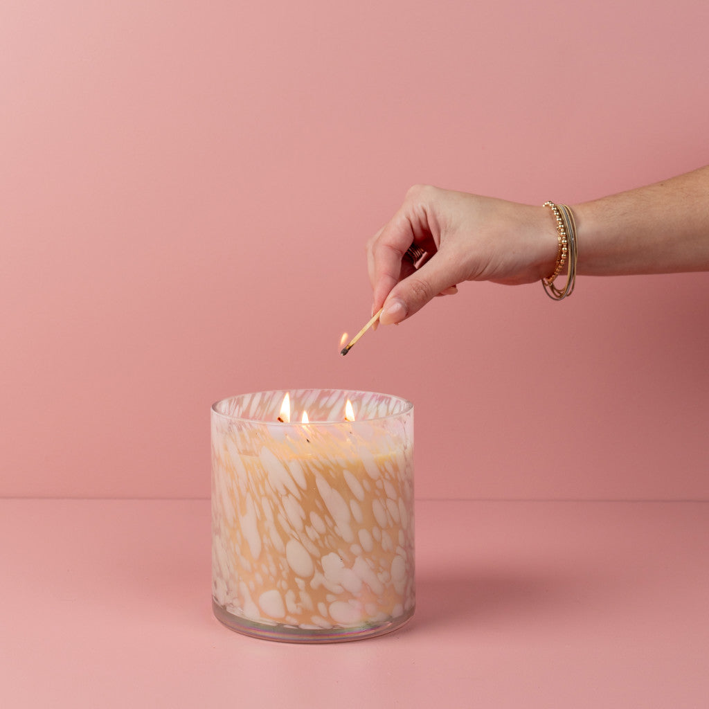 A hand with bracelets lights the Sweet Grace Collection Candle #058 by Bridgewater Candle Company, a large cream marbled three-wick soy-blend candle, using a matchstick against a soft pink background.