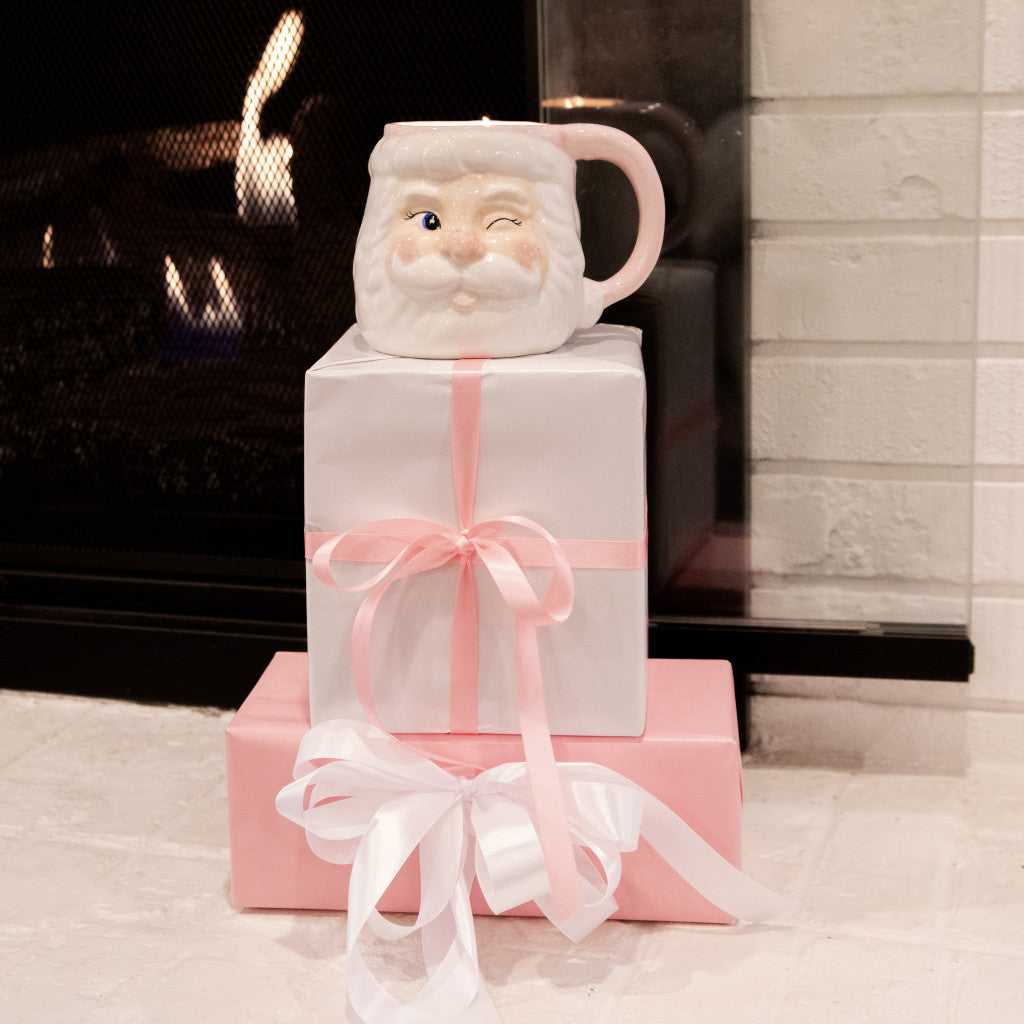 A winking Bridgewater Candle Company Sweet Grace Santa Mug sits atop two wrapped gifts by the fireplace. Enjoy its festive scent, then reuse the Santa Mug for holiday cheer all season from Bridgewater Candle Company.
