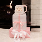 A winking Bridgewater Candle Company Sweet Grace Santa Mug sits atop two wrapped gifts by the fireplace. Enjoy its festive scent, then reuse the Santa Mug for holiday cheer all season from Bridgewater Candle Company.