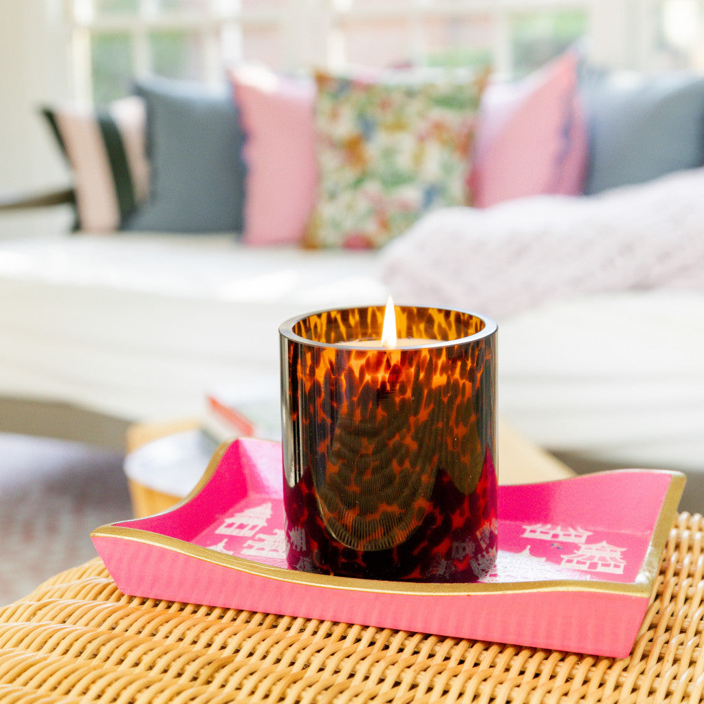 A Bridgewater Candle Company Sweet Grace Collection Candle #064, featuring soy-blend wax in a brown tortoise glass, rests on a pink tray atop a wicker table with blurred colorful pillows and a sofa in the background.