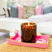 A Bridgewater Candle Company Sweet Grace Collection Candle #064, featuring soy-blend wax in a brown tortoise glass, rests on a pink tray atop a wicker table with blurred colorful pillows and a sofa in the background.