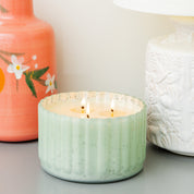 A Bridgewater Candle Company Sweet Grace Collection Candle #065, featuring an antique green ribbed vessel with three wicks and soy-blend wax, sits on a gray surface between a pink floral vase and a white textured lamp.