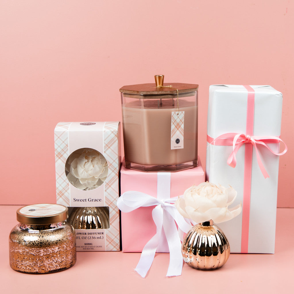 A collection of pink and white gifts, featuring the Bridgewater Candle Company Sweet Grace Mercury Flower Diffuser and candles, arranged with wrapped boxes—perfect for adding a delightful home fragrance to any space.