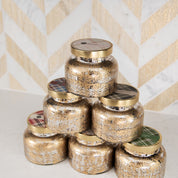 Six Bridgewater Candle Company Sweet Grace Mercury Jar Candles, featuring gold-speckled glass jars with metallic lids, are stacked in a pyramid on a white surface before a chevron-patterned backdrop of white, gold, and gray tiles.