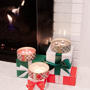 Three lit Christmas Bliss Holiday 3-Wick Tin Candles by Bridgewater Candle Company in plaid tins sit atop gift-wrapped boxes with red and green ribbons, arranged in front of a glowing fireplace with a white brick surround.