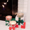 Three lit Christmas Bliss Holiday 3-Wick Tin Candles by Bridgewater Candle Company in plaid tins sit atop gift-wrapped boxes with red and green ribbons, arranged in front of a glowing fireplace with a white brick surround.