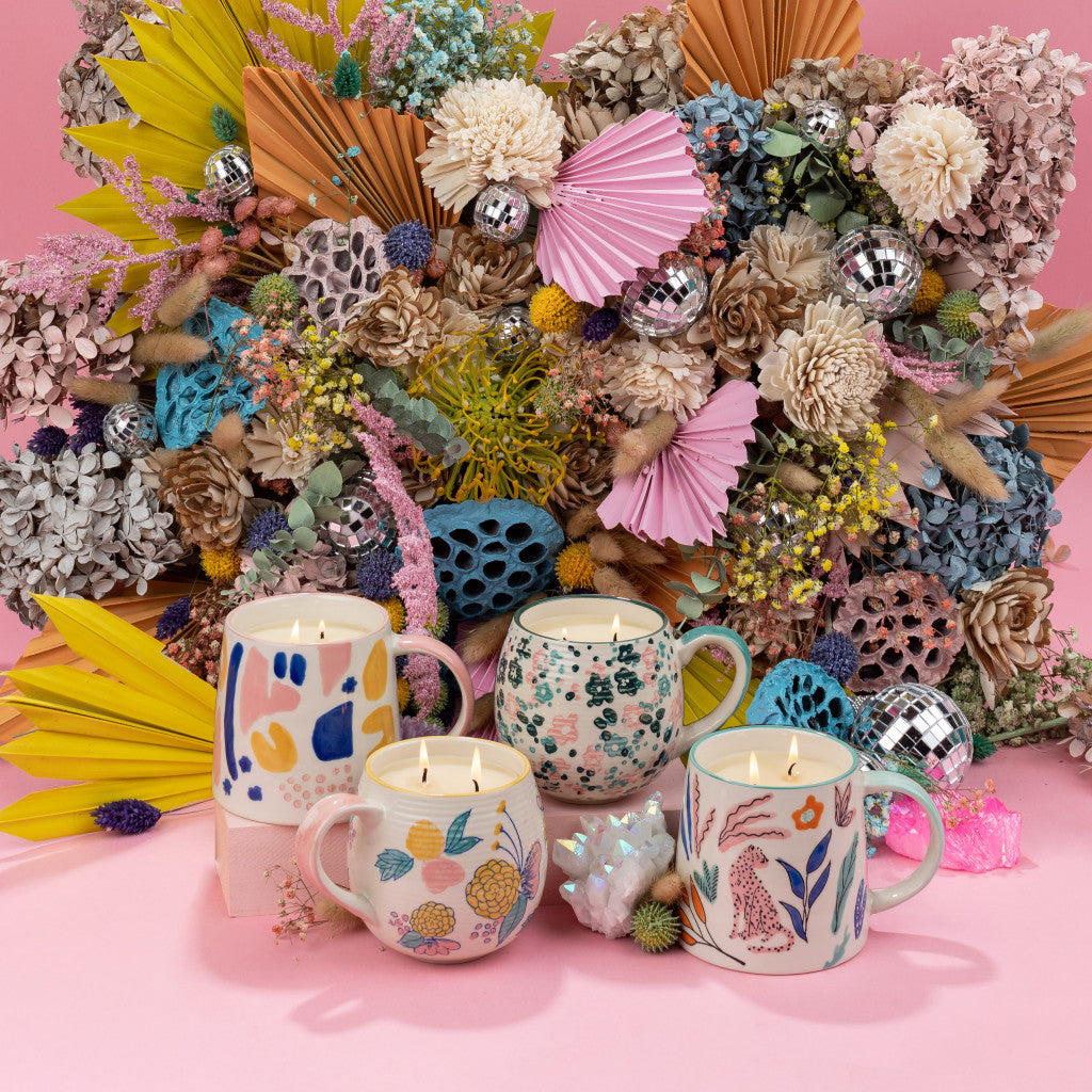 A vibrant floral bouquet of dried and artificial blooms sits behind four Bridgewater Candle Company Sweet Grace Collection Candle #048 soy-blend candles in patterned mugs on a pink surface, surrounded by disco balls and crystals.