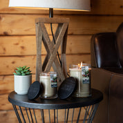 A round black side table holds two Afternoon Retreat 8oz Jar Candles by Bridgewater Candle Company—one lit, one unlit—each with a black lid, plus a potted succulent and wooden lamp, set by wood-paneled walls near a brown leather chair.
