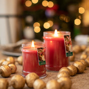 Two Christmas Bliss 18oz Large Jars by Bridgewater Candle Company with glowing flames sit on a table with gold beads, while a Christmas tree shines behind them, filling the room with a festive, warm holiday fragrance.