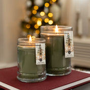 Two lit Bridgewater Candle Company Festive Frasier 18oz Large Jar Candles, each with a festive pine tree label, sit on a closed book with blurred holiday lights and decorations in the background.