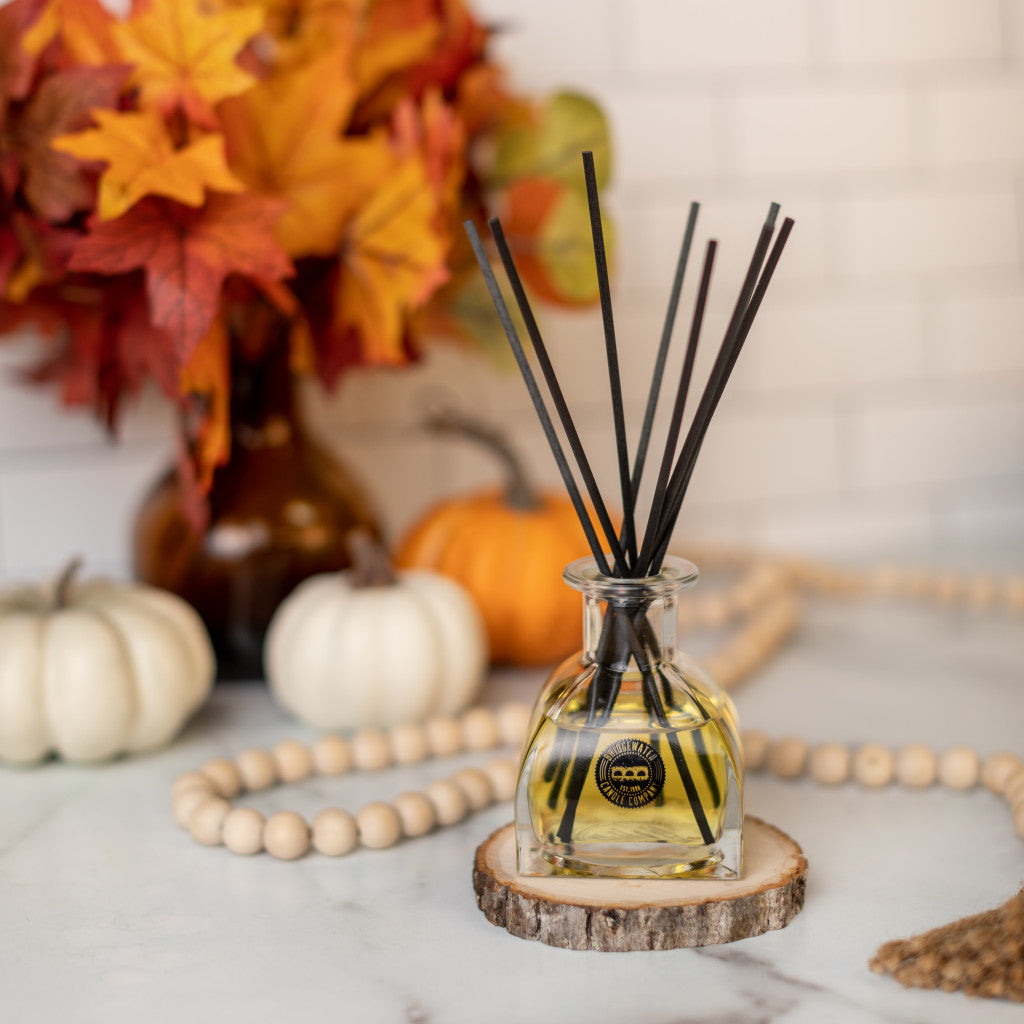 A Bridgewater Candle Company Harvest Pumpkin Reed Diffuser with black reeds sits on a wooden coaster, surrounded by beads, pumpkins, and autumn leaves—bringing cozy fall fragrance to your marble surface.