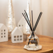 A Festive Frasier Reed Diffuser by Bridgewater Candle Company, with black fiber reeds, is displayed on wood among white decorative houses and silver cones, infusing the area with its signature holiday fragrance.