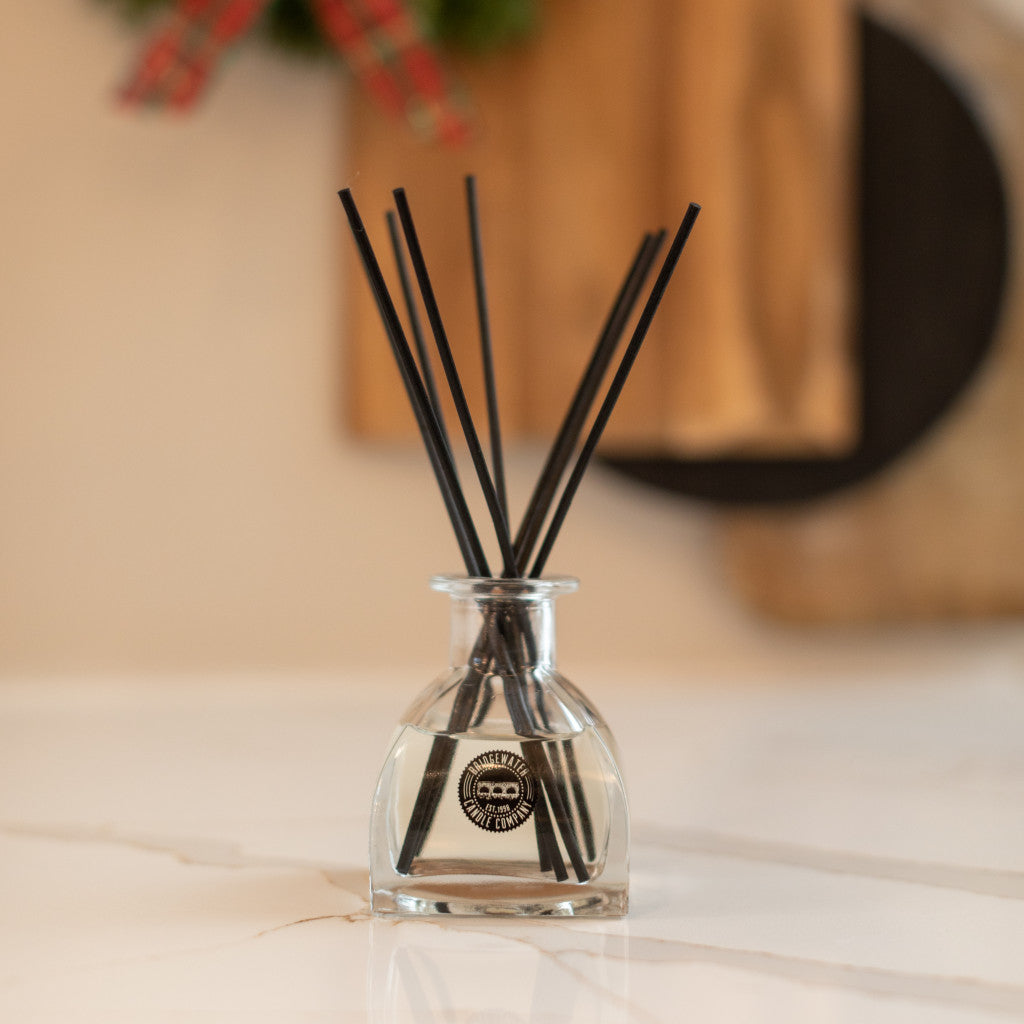 A clear glass Afternoon Retreat Reed Diffuser by Bridgewater Candle Company, filled with fragrance and black fiber reeds, stands on a white surface. Behind it are soft brown tones, greenery, and a touch of red ribbon in the blurred background.