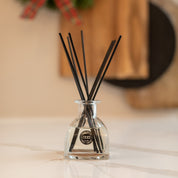 A clear glass Afternoon Retreat Reed Diffuser by Bridgewater Candle Company, filled with fragrance and black fiber reeds, stands on a white surface. Behind it are soft brown tones, greenery, and a touch of red ribbon in the blurred background.