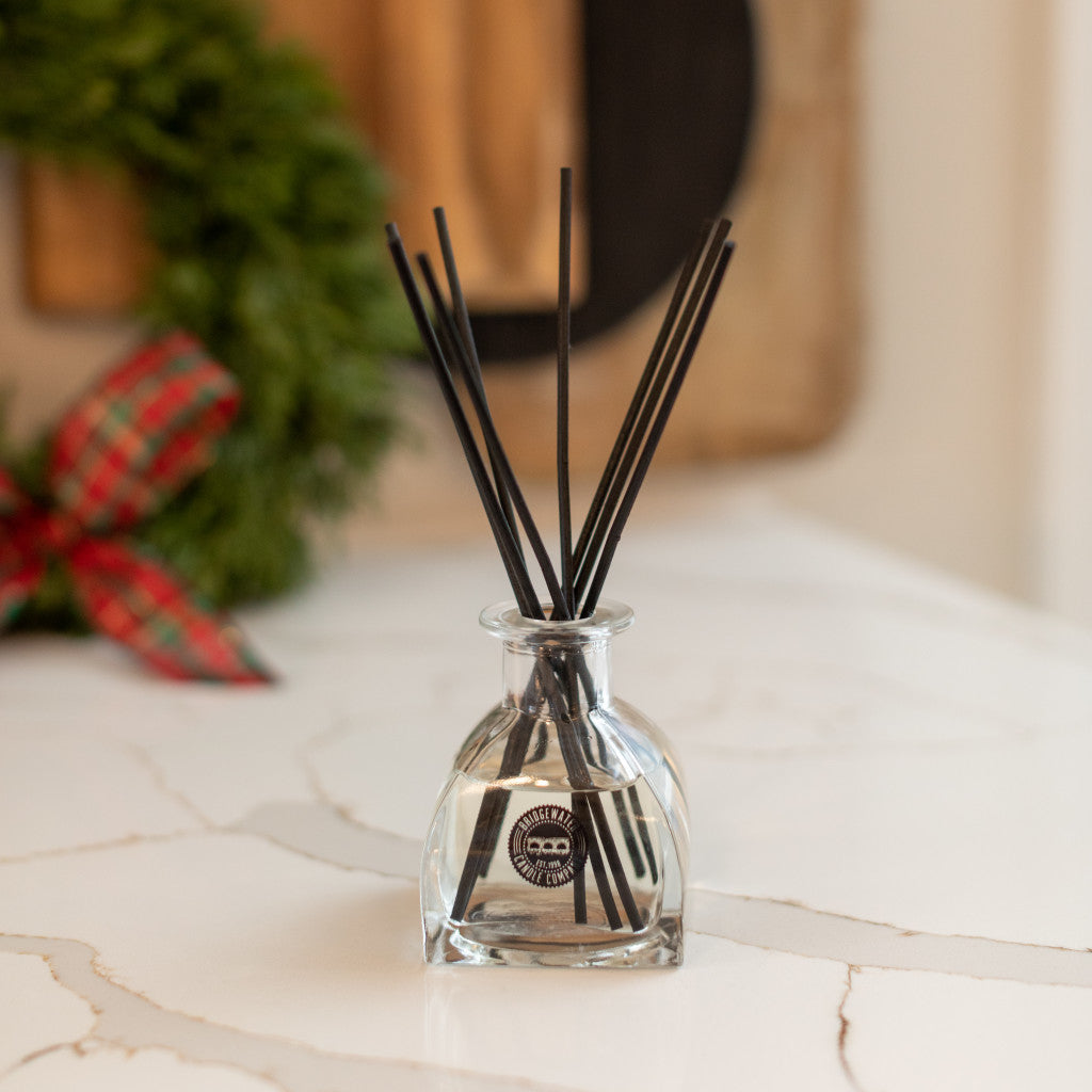 A clear Christmas Bliss Reed Diffuser by Bridgewater Candle Company with black reeds rests on white marble, while a green wreath with a red and green plaid bow adds festive cheer in the background.