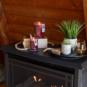 A cozy scene featuring the Bridgewater Candle Company Welcome Home 8oz Jar Candle, two glowing red candles on stacked books, faux white-potted plants, a string of wooden beads, and a gold candle holder atop a dark mantel against wood paneling.