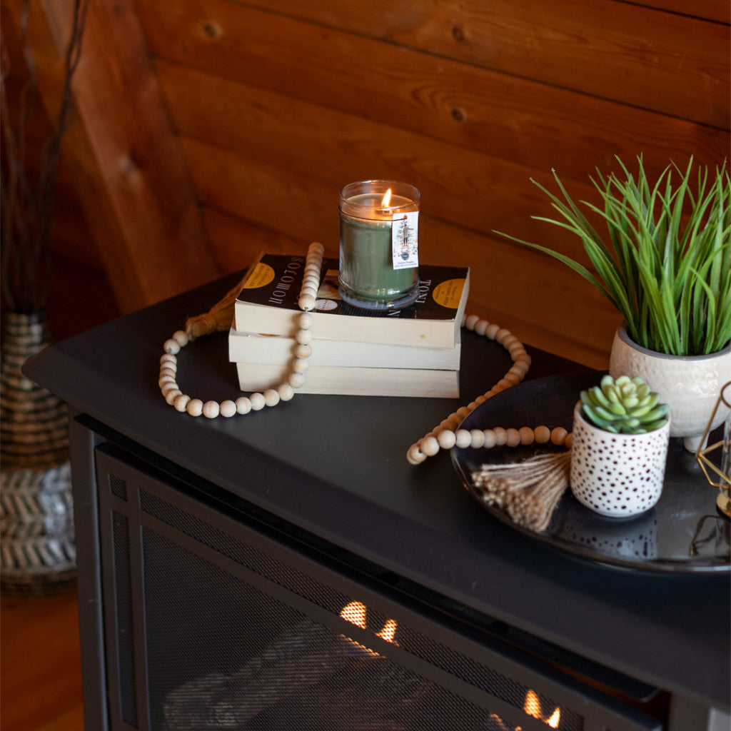 A lit Bridgewater Candle Company Festive Frasier 8oz Jar Candle sits on stacked books with a wooden bead garland, beside potted plants and a decorative tray on a dark mantel in a cozy, wood-paneled room.