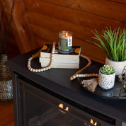 A lit Bridgewater Candle Company Festive Frasier 8oz Jar Candle sits on stacked books with a wooden bead garland, beside potted plants and a decorative tray on a dark mantel in a cozy, wood-paneled room.