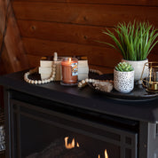 A cozy scene with a Bridgewater Candle Company Harvest Pumpkin 8oz Jar Candle glowing amid stacked books, a wooden bead garland, potted plants, and small decor on a black fireplace mantel. Warm fire below; wood-paneled wall behind.