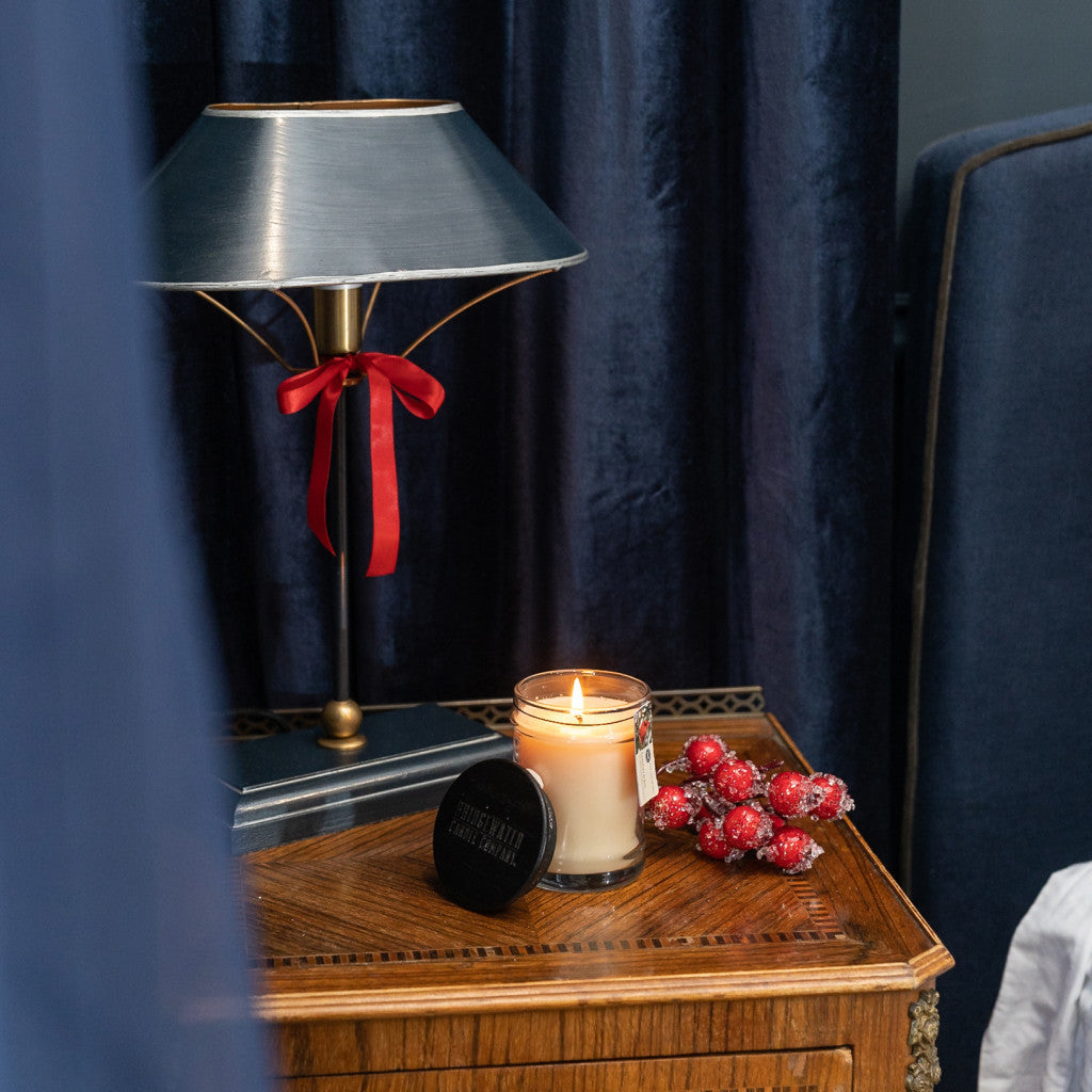 A Bridgewater Candle Company Comfort & Joy 8oz Small Jar Candle, red sugared berries, and a black tin sit on a wooden nightstand by a lamp with a red ribbon, adding warmth and festivity against dark blue drapes and furniture.