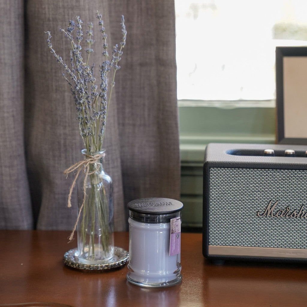 A small glass vase with dried lavender, a Bridgewater Candle Company Lavender Fields 8oz Small Jar Candle, and a vintage-style speaker sit on a wooden surface in front of a curtained window.
