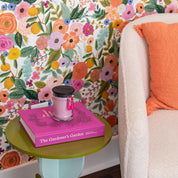 A cozy nook features a textured ivory chair with an orange pillow, a green side table holding Bridgewater Candle Companys XOXO 8oz Small Jar Candle, and the pink book The Gardener’s Garden, all set against vibrant floral wallpaper.