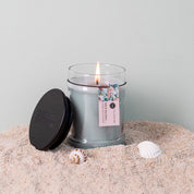 A Bridgewater Candle Company Salt & Sea Glass 8oz Small Jar Candle, made with soy-blend wax, sits on sand next to three seashells in a glass jar with a black lid against a pale background.