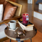 A small round wooden side table holds a ceramic turtle, a gray mug on a coaster, two stacked books, and a lit Bridgewater Candle Company Welcome Home 18oz Large Jar Candle. A wooden chair with tan cushions and stairs are in the background.