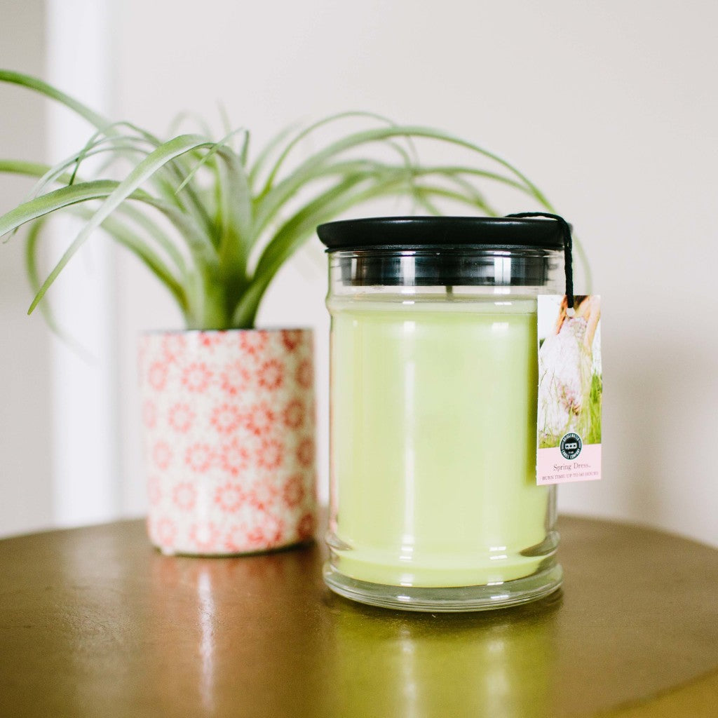 The Spring Dress 18oz Large Jar Candle by Bridgewater Candle Company in green sits in a clear glass jar with a black lid on wood beside a potted plant, bringing Bridgewater fragrance to your space.