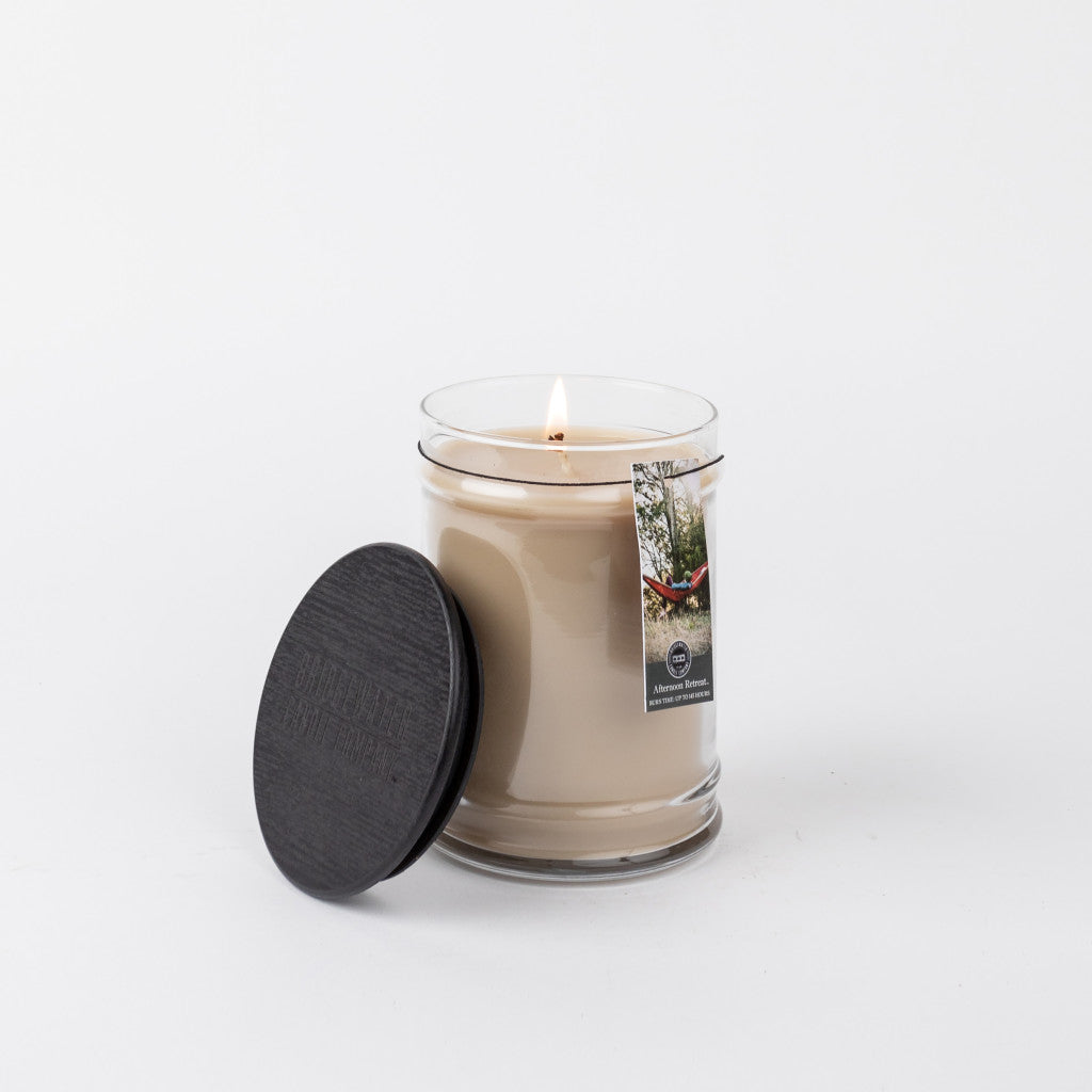A lit Bridgewater Candle Company Afternoon Retreat 18oz Large Jar Candle in clear glass with a black lid beside it. The label shows a red hammock among trees, set against a plain white background.