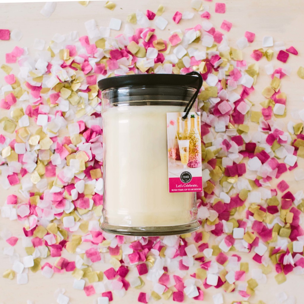 The Lets Celebrate 18oz Large Jar Candle by Bridgewater Candle Company, in a glass jar with a black lid and tag, sits on confetti, filling the air with its delightful fragrance.