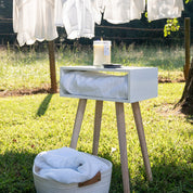 Outside on the grass, a white bedside table holds a Bridgewater Candle Company Laundry Line 18oz Large Jar Candle on top and a folded towel inside. In the background, white clothes hang on a line with a basket of towels nearby in the sunlight.