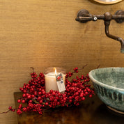 A glowing Comfort & Joy 18oz Large Jar Candle by Bridgewater Candle Company, surrounded by red berries, sits on a dark wood surface near a green ceramic sink and rustic faucet, adding warmth to the cozy setting.