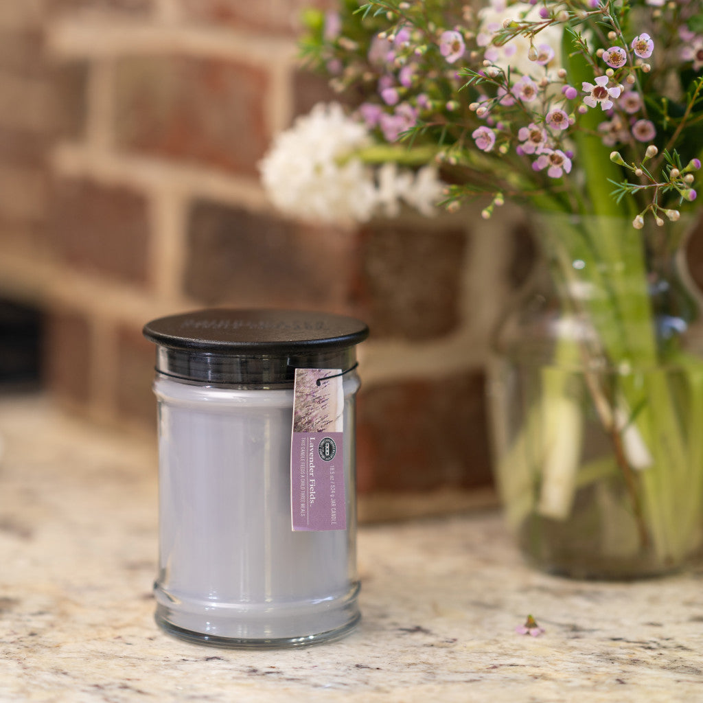 A Bridgewater Candle Company Lavender Fields 18oz Large Jar Candle with a black lid rests on a marble countertop beside a glass vase of white and pink flowers, set against a brick wall backdrop.