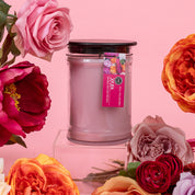The XOXO 18oz Large Jar Candle by Bridgewater Candle Company features soy-blend wax in a glass jar with a dark lid, displayed among colorful artificial roses and peonies against a pink background—perfect for any occasion.