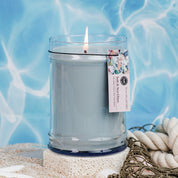 The Bridgewater Candle Company Salt & Sea Glass 18 oz Large Jar Candle features soy-blend wax in a clear glass jar with a floral label, displayed amid seashells and rope on sand against a blue, ocean-like background.