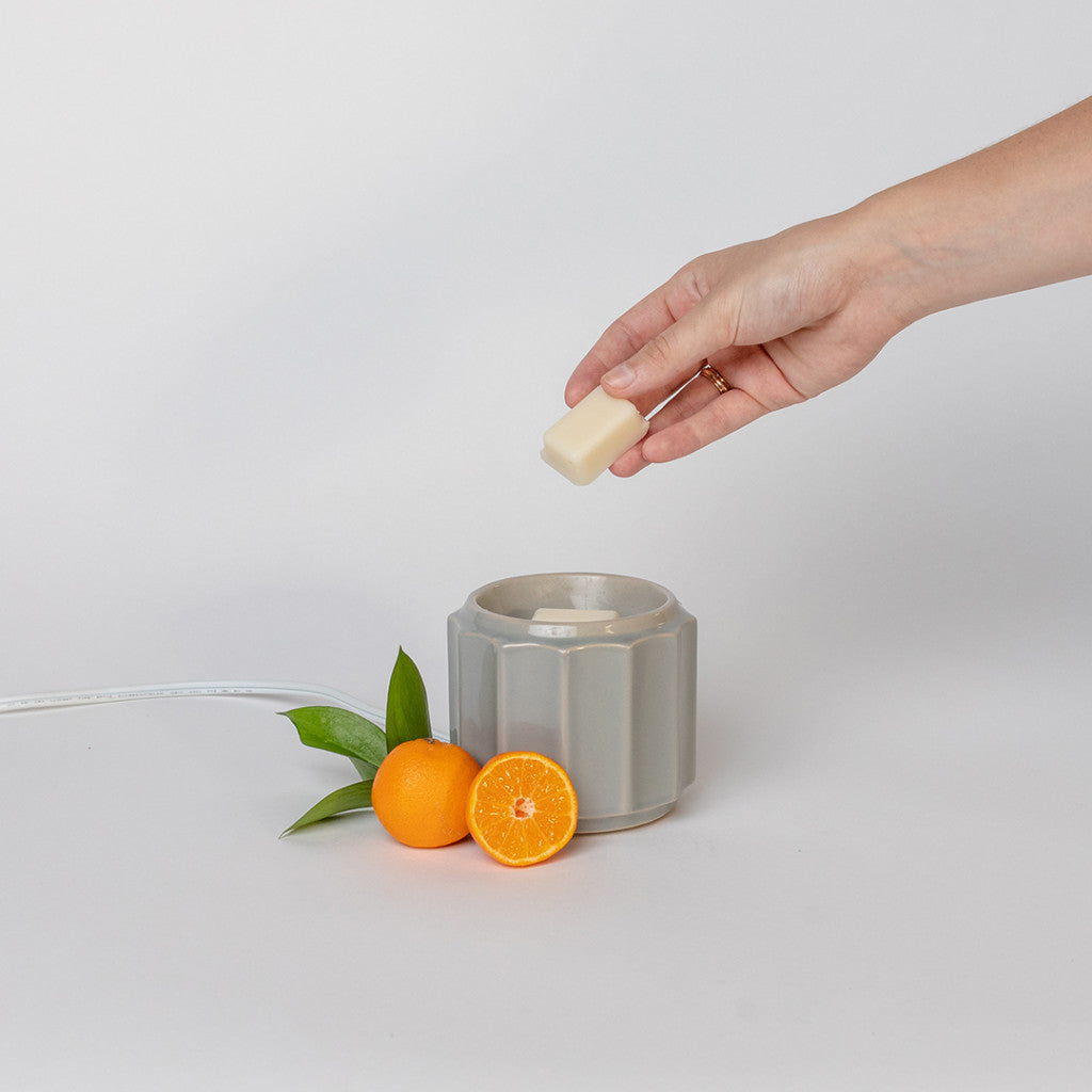 A hand places a wax melt into the Bridgewater Candle Company Wax Warmer-Grey Banded, with a whole and halved orange plus green leaves beside it on a white background.