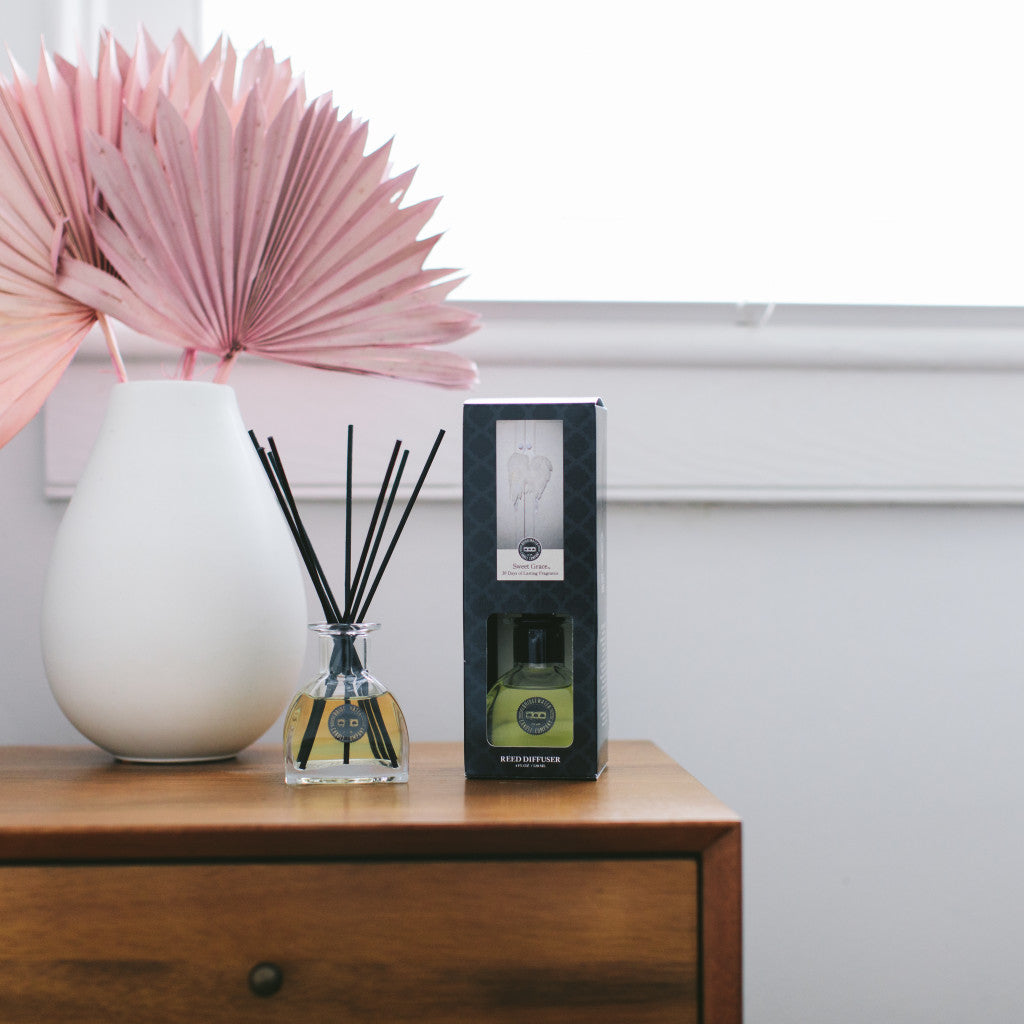 A Bridgewater Candle Company Sweet Grace Reed Diffuser and its box rest on a wooden nightstand beside a white vase with pink dried palm leaves, creating a chic home fragrance display against the white wall and window.
