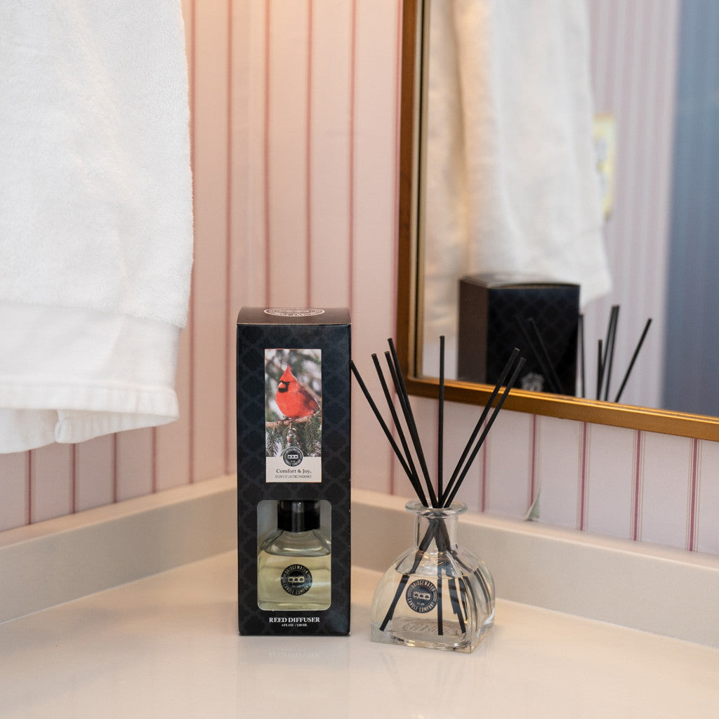 A glass reed diffuser and a boxed diffuser set with a cardinal image sit on a bathroom countertop next to a mirror and a white towel hanging on the wall.