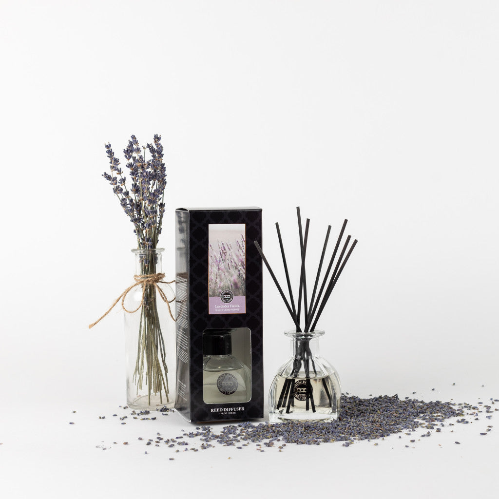 A glass bottle with dried lavender stems, a boxed Bridgewater Candle Company Lavender Fields Reed Diffuser, and a glass diffuser bottle with black fiber reeds are displayed among scattered lavender buds on a white background.