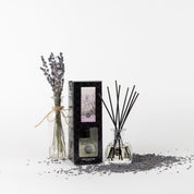 A glass bottle with dried lavender stems, a boxed Bridgewater Candle Company Lavender Fields Reed Diffuser, and a glass diffuser bottle with black fiber reeds are displayed among scattered lavender buds on a white background.