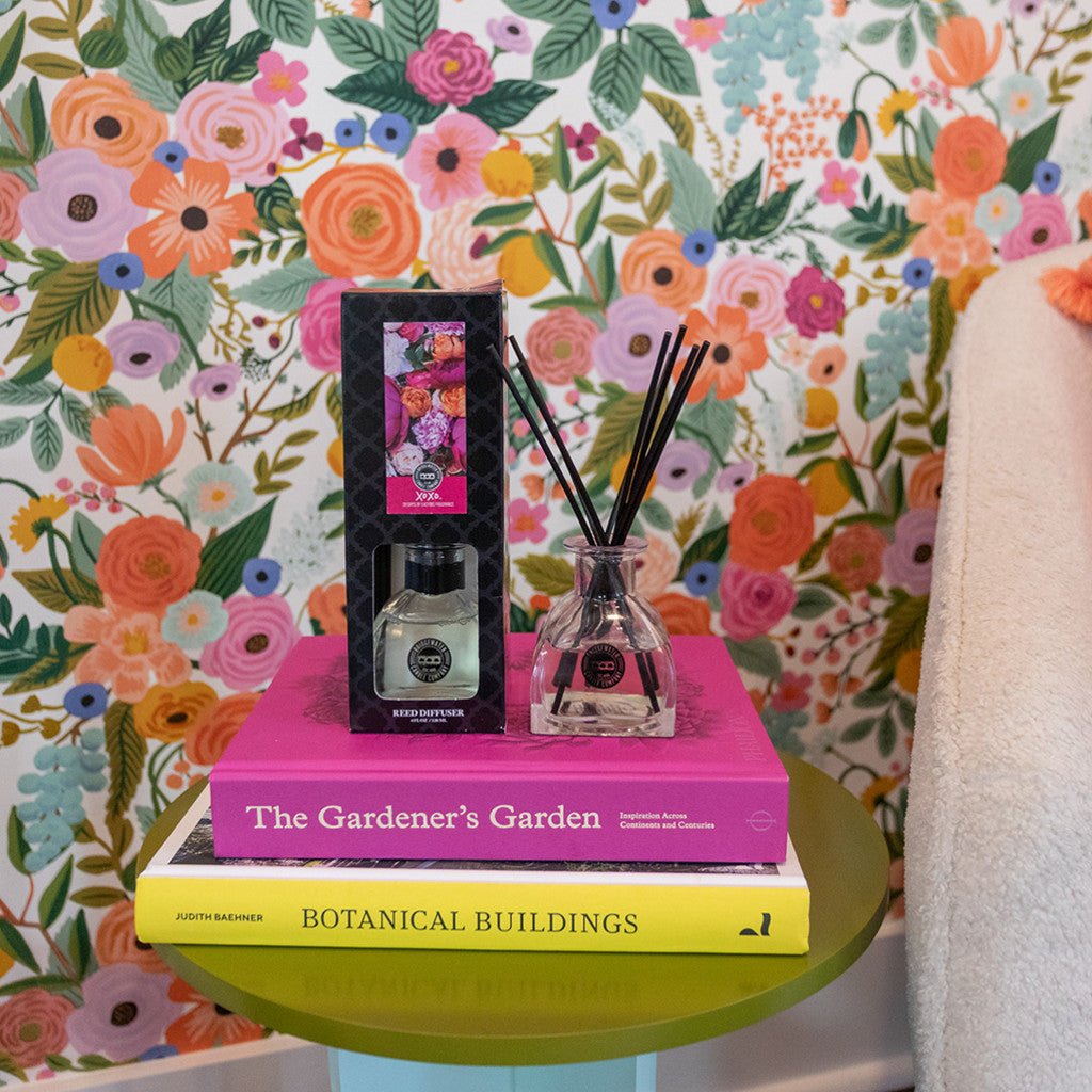 The XOXO Reed Diffuser by Bridgewater Candle Company, featuring fiber reeds and floral packaging, is displayed on The Gardener’s Garden and Botanical Buildings books atop a green table with vibrant floral wallpaper in the background.