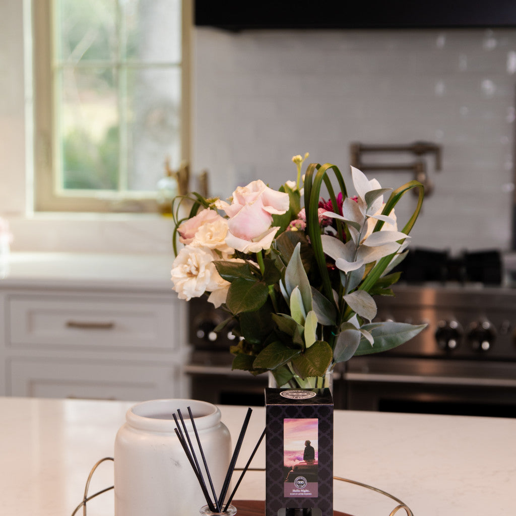 A vase of assorted flowers and a Bridgewater Candle Company Hello Night Reed Diffuser with fiber reeds sit on a tray on the kitchen counter, adding premium fragrance to the space against white cabinets and a window backdrop.