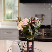 A vase of assorted flowers and a Bridgewater Candle Company Hello Night Reed Diffuser with fiber reeds sit on a tray on the kitchen counter, adding premium fragrance to the space against white cabinets and a window backdrop.