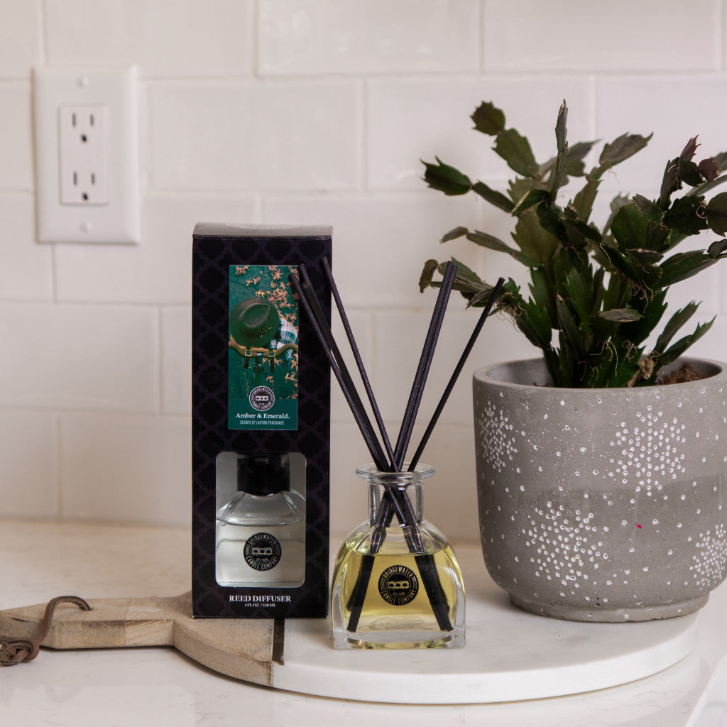 A Bridgewater Candle Company Amber & Emerald Reed Diffuser with fiber reeds and premium fragrance oil in a glass bottle sits on a white counter next to its box and a speckled gray potted plant, against a white tiled wall.