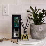 A Bridgewater Candle Company Amber & Emerald Reed Diffuser with fiber reeds and premium fragrance oil in a glass bottle sits on a white counter next to its box and a speckled gray potted plant, against a white tiled wall.